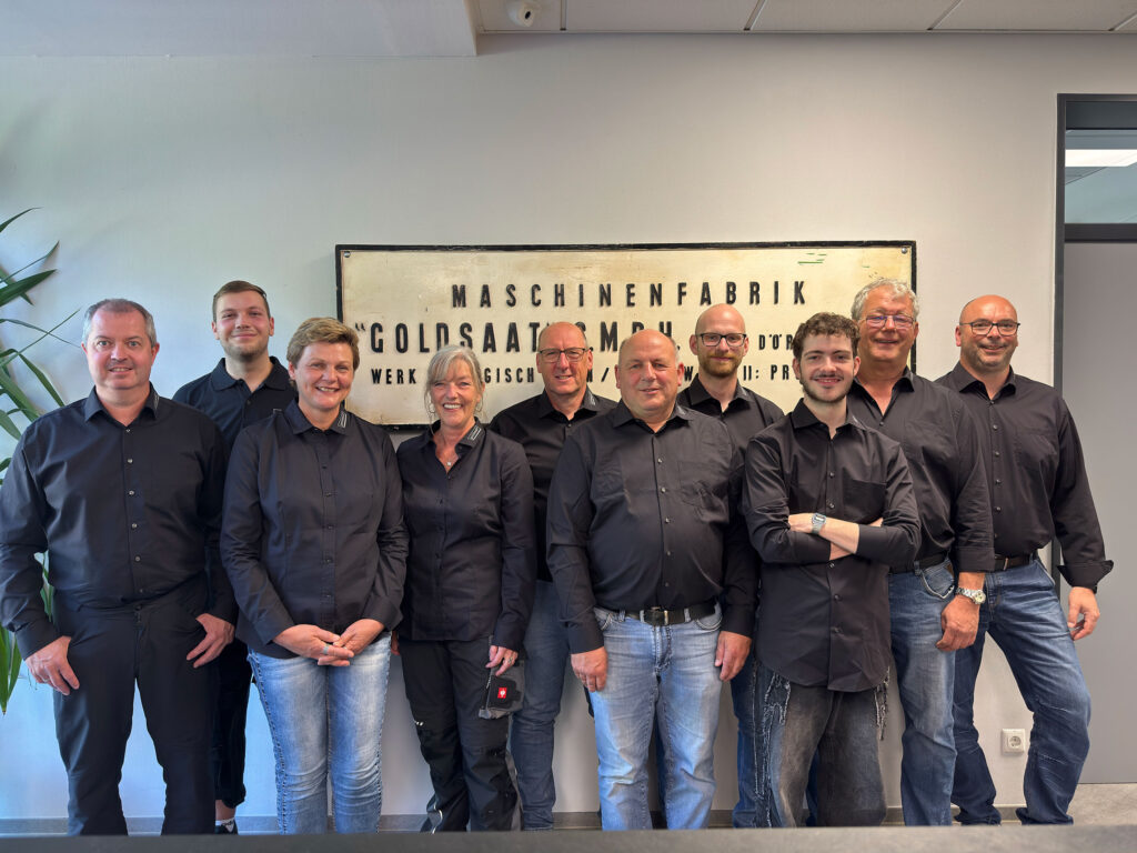Das Büro-Team von Goldsaat Agrartechnik steht gemeinsam für ein Gruppenfoto vor einer Wand mit historischem Firmenschild und lächelt in die Kamera.