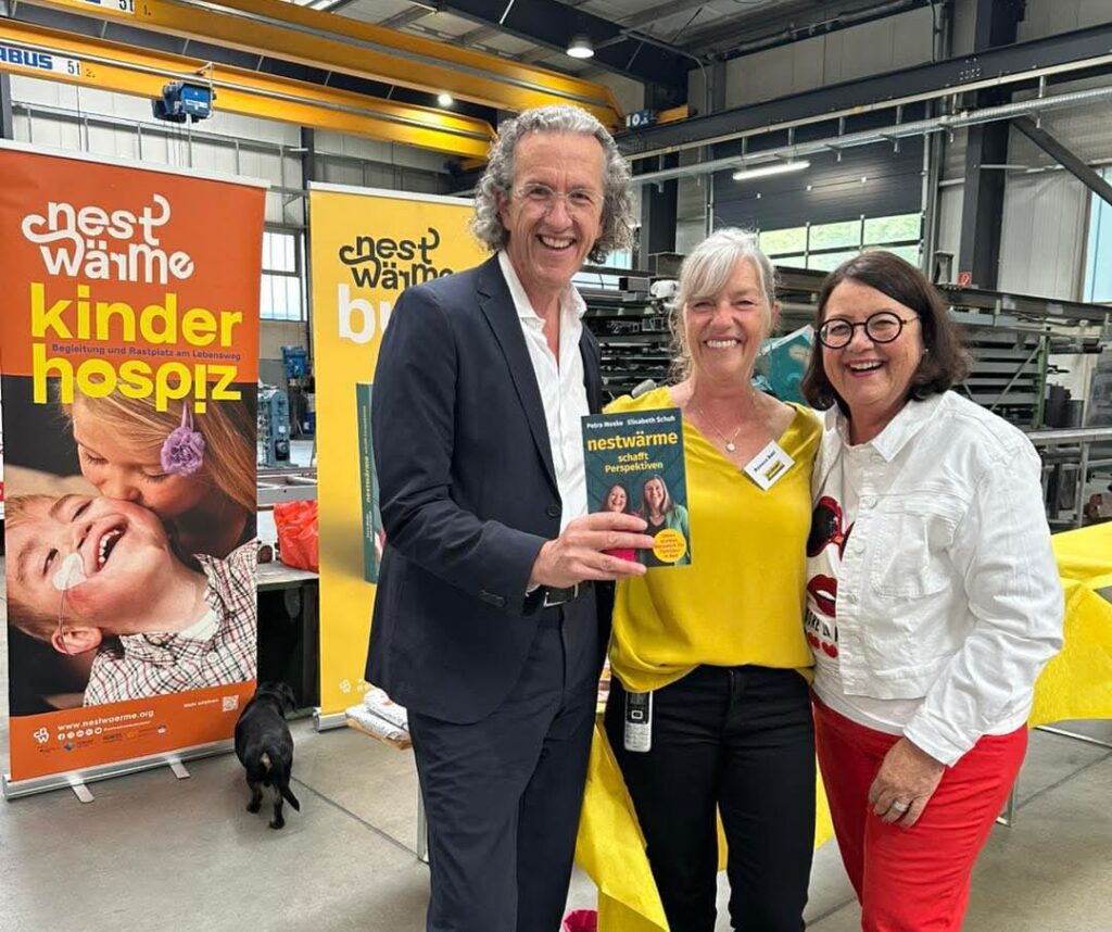 Drei lachende Personen stehen bei einer Spendenaktion zugunsten eines Kinderhospizes in einer Werkhalle und halten ein Buch mit dem Titel ‚Nestwärme schafft Perspektiven‘.